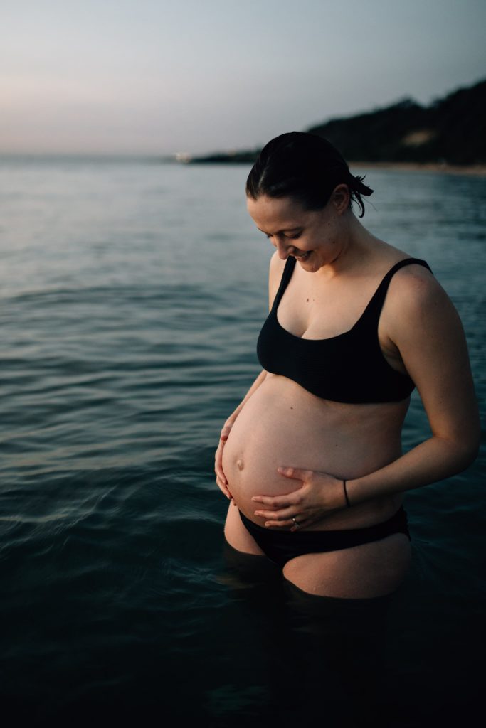 woman in black bikini looking at pregnant belly during beach maternity photos, And So I Don't Forget Photography