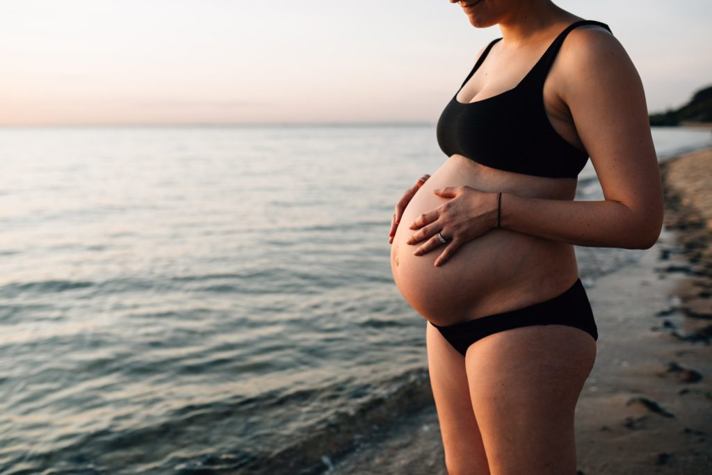 woman holding pregnant belly on shore duuring beach maternity photos, And So I Don't Forget Photography