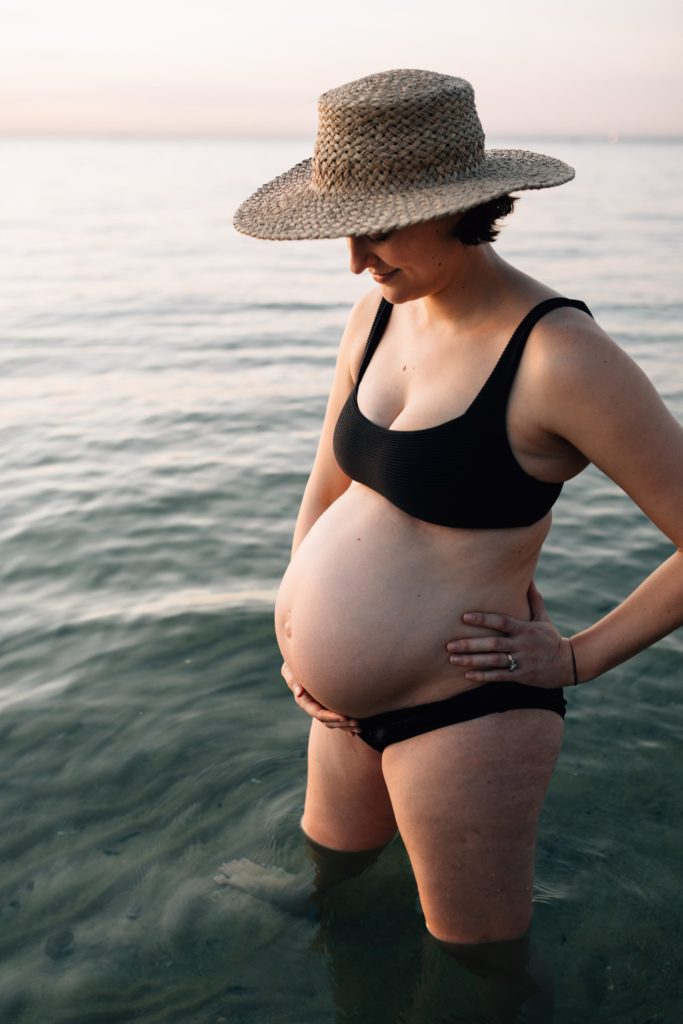 woman standing in water wearing bikini and hat during beach maternity photos, And So I Don't Forget Photography