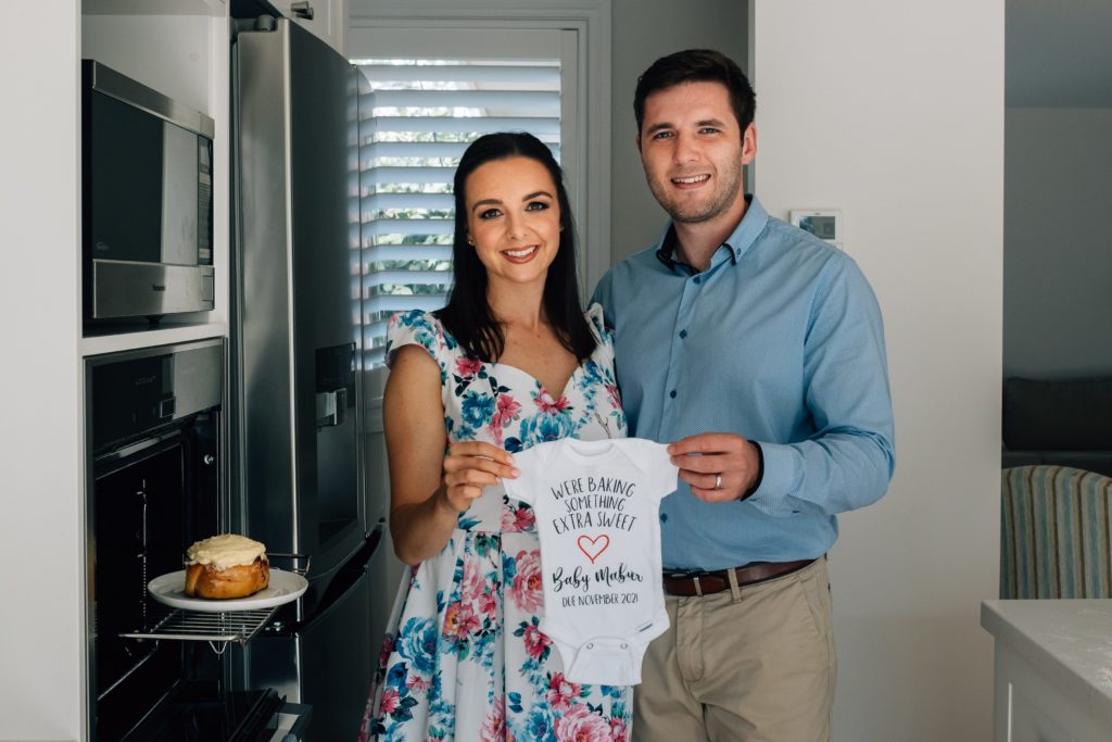 husband and wife smiling at camera in kitchen with bun in the oven pregnancy announcement photography session