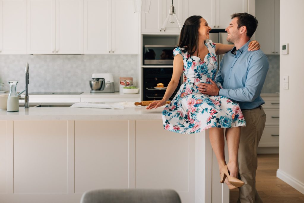 bun in the oven in background with man and woman in kitchen laughing, funny pregnancy announcement ideas 