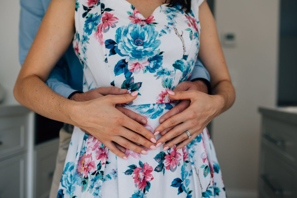 two sets of hands on belly of woman in floral dress, bun in the oven pregnancy announcement photography session
