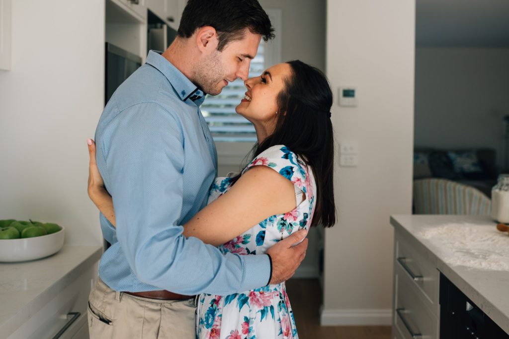 husband and wife brushing noses together in kitchen during bun in the oven pregnancy announcement photography session