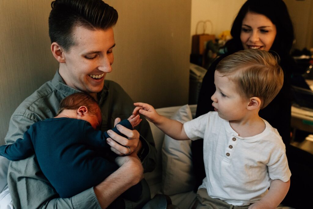 Dad holding baby and smiling, son touching baby's hand and Mum smiling in background watching. Fresh 48 newborn session