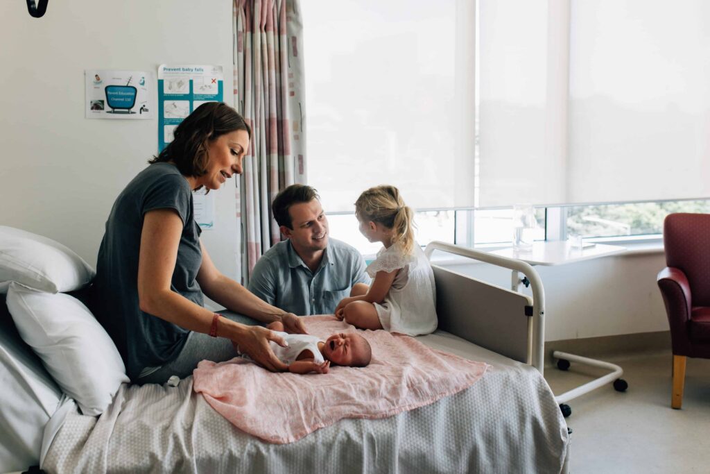 Girl sitting on hospital bed while meeting newborn sibling