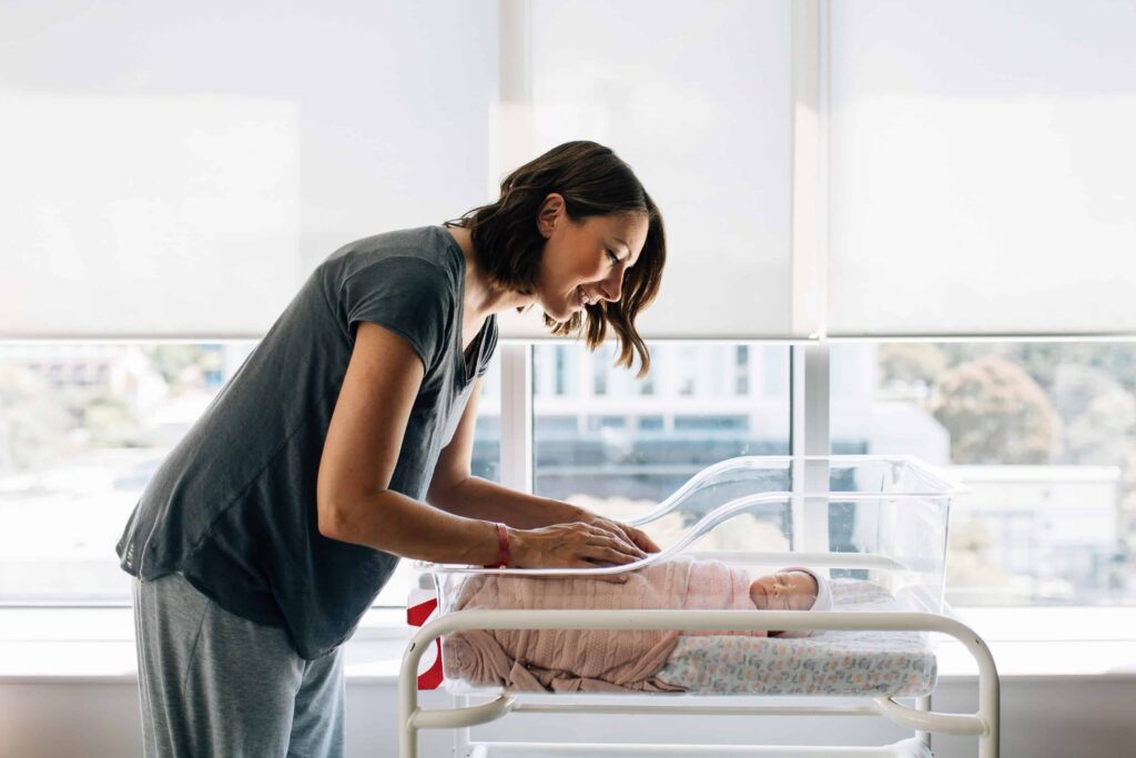 Mum leaning over newborn baby's bassinette in front of hospital windows