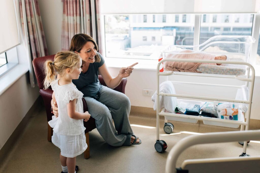 Mum pointing out newborn baby to her big sister during their first meeting